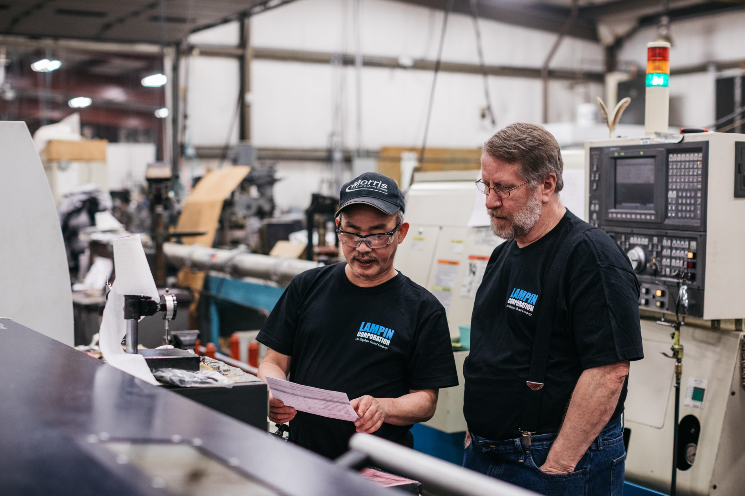 Two employee-owners at Lampin discuss a work document. Both are wearing black Lampin tshirts, one is holding a pink sheet of paper.