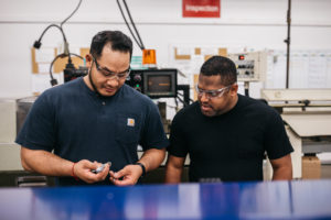 Two of Lampin's employee owners engaged in conversation about on-the-job cross-training. One wears a blue tshirt and holds an inspection gauge; the other is wearing a black tshirt and looking at the gauge in his fellow ESOP participant's hand.