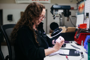 Another Lampin ESOP participant and employee-owner inspects a part for deburring. She has long, curly, dark hair and a black shirt. She holds a small metal piece under an illuminated magnifier.