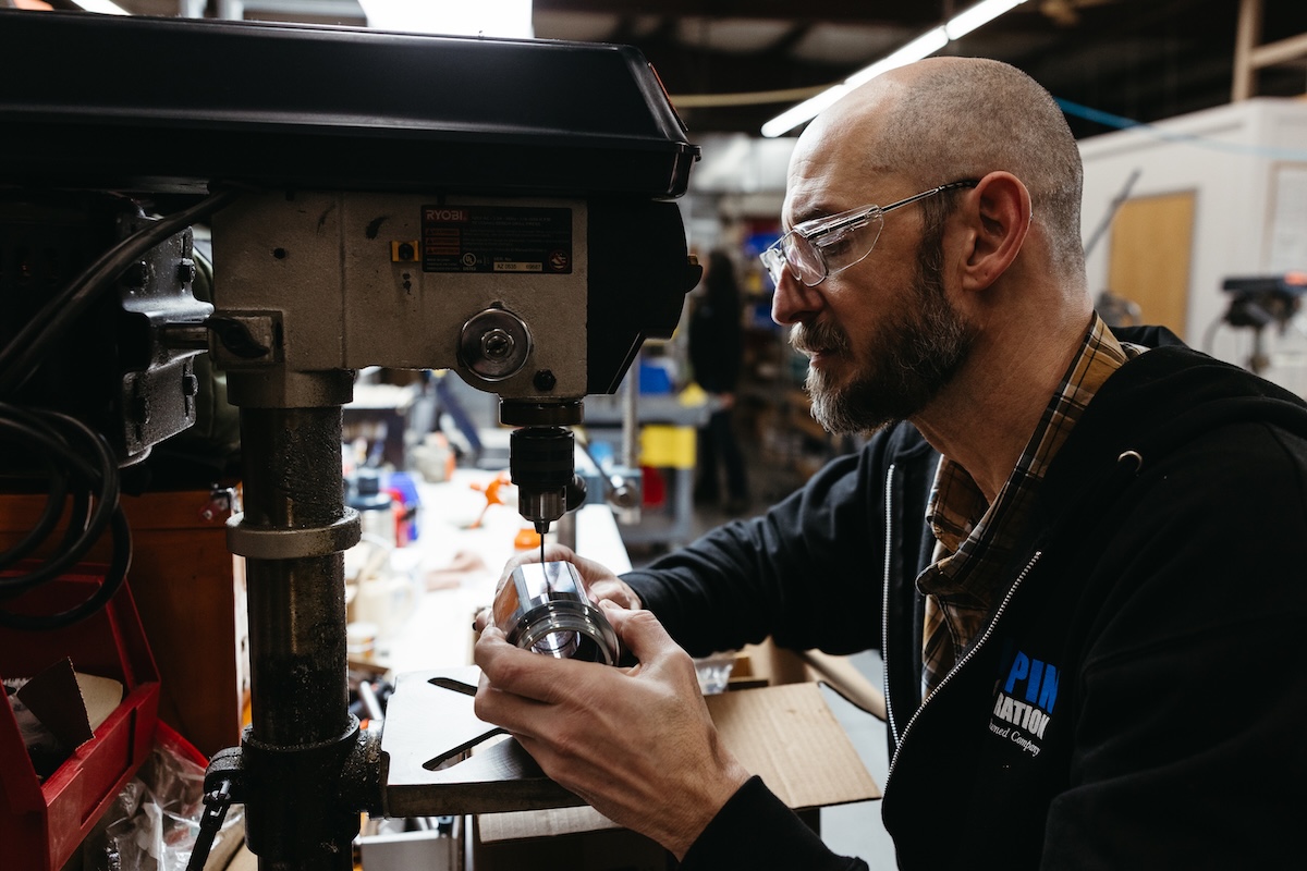 One of Lampin's employee owners, wearing safety glasses and a Lampin Corporation hooded sweatshirt, uses a small benchtop tool to drill into a part.